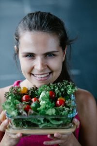 A happy woman in a pink top holds a healthy vegetable salad, showcasing fresh greens and cherry tomatoes.