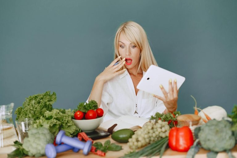 A surprised woman looking at a tablet surrounded by fresh vegetables and fruits, promoting healthy eating.