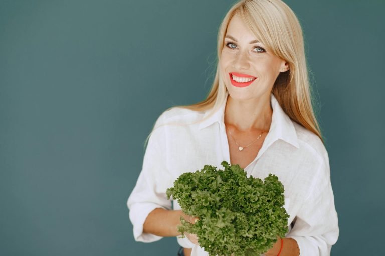 Cheerful woman with blonde hair holding fresh lettuce, promoting healthy living in a studio setting.